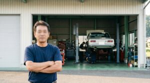 岡山県の地方都市の小さな車屋の前で、50代の男性経営者が腕組みをして店の前に立っている。整備工場の入口、整備中の車が見える、午前の柔らかい光、写真風、顔はぼかす