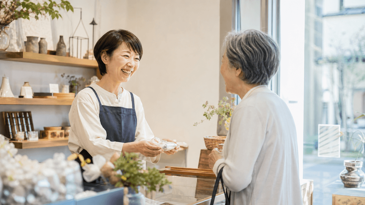 日本人の40代女性の小規模事業者が、明るい店内で常連客らしき年配の女性と笑顔で会話している場面。信頼関係が感じられる温かい雰囲気。写真風、ドキュメンタリーテイスト、自然光