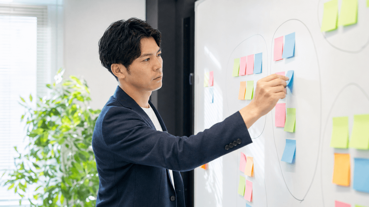 日本人の30代男性経営者が、オフィスのホワイトボードに顧客リストを書き出し、付箋で分類しながら整理している場面。戦略的に考えている様子。写真風、ドキュメンタリーテイスト、明るいオフィス