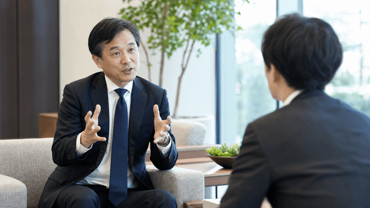 日本人の50代男性経営者が、聞き手(インタビュアー)に向かって身振りを交えながら熱心に語っている場面。対話している様子。明るい応接室、自然光、写真風、ドキュメンタリーテイスト