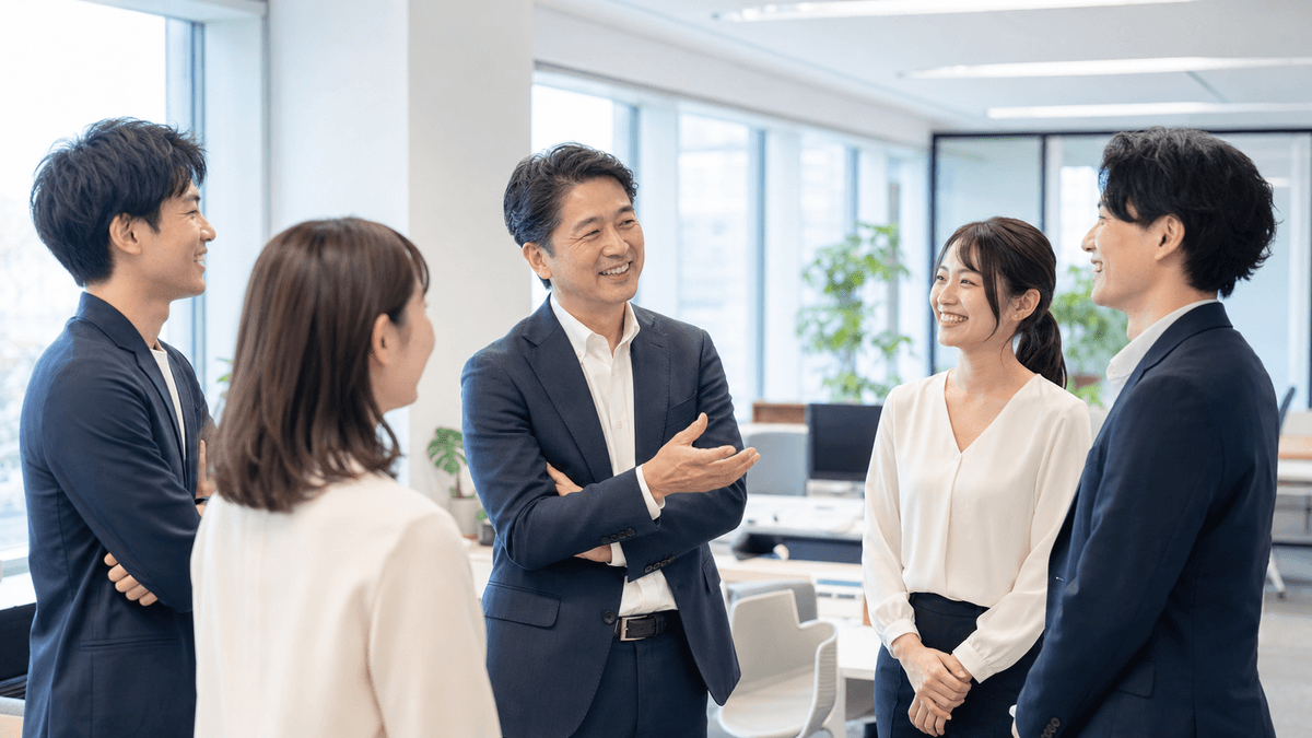 日本人の経営者が、明るいオフィスで社員たちと自然な笑顔で立ち話をしている場面。緊張感のない、信頼関係のある職場の雰囲気。明るい自然光、写真風、前向きで温かいトーン
