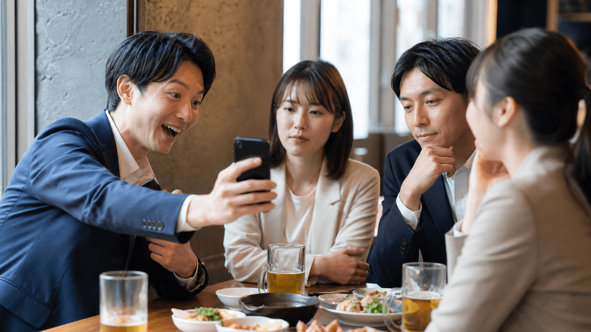 日本人のビジネスパーソンたちが居酒屋やオフィスで談笑している場面。一人がスマホを見せて「AIすごいよね」と盛り上げているが、聞いている側はどこか他人事のような表情。熱量の差を感じさせる構図、写真風、自然光、リアルな雰囲気