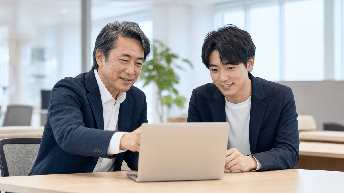 日本人の経営者と若手社員が、オフィスで一台のパソコンを一緒にのぞき込み、小さな作業を試している場面。肩の力が抜けた、前向きで和やかな雰囲気。明るい自然光、写真風、ドキュメンタリーテイスト