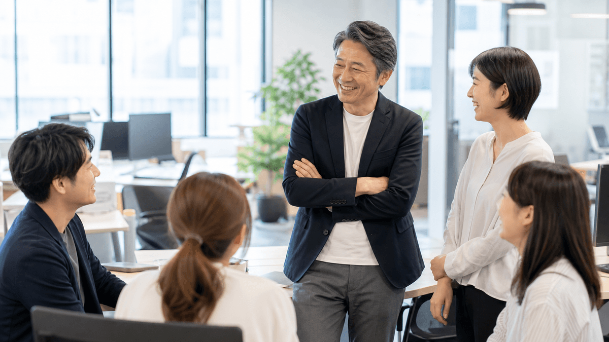 日本人の経営者が、明るいオフィスで社員たちと自然な笑顔で立ち話をしている場面。緊張感のない、信頼関係のある職場の雰囲気。明るい自然光、写真風、前向きで温かいトーン