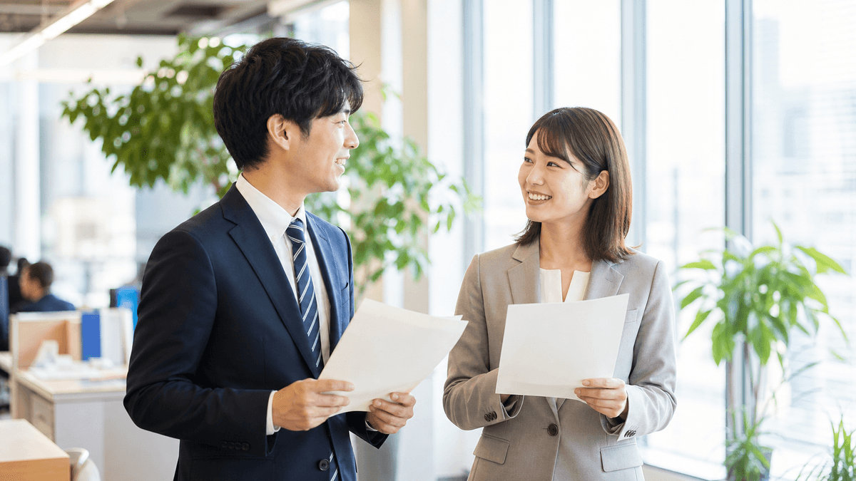 日本人の20代から30代の若いビジネスパーソン男女2人が、明るいオフィスで前向きな表情で資料を手に立ち話をしている場面。窓から差し込む朝の光、希望のある雰囲気。写真風、ドキュメンタリーテイスト